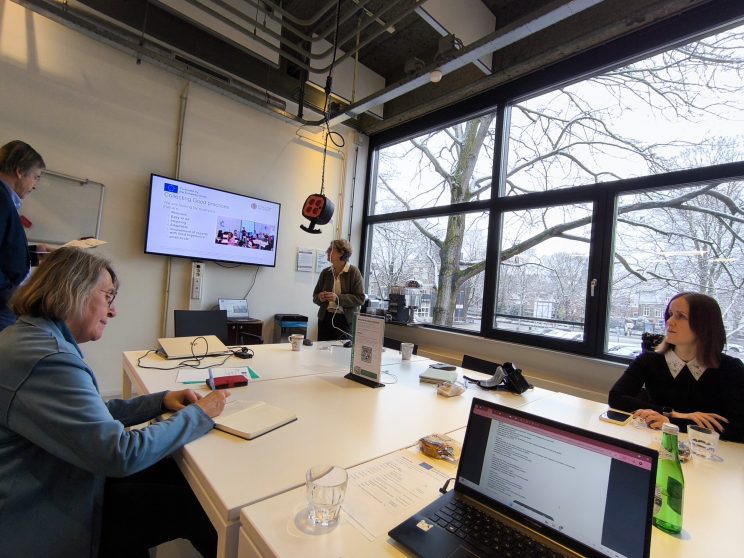 Na fotografiji je vidljivo pet osoba koje sudjeluju na sastanku u modernoj konferencijskoj sobi s velikim prozorima kroz koje se vidi snježan krajolik. Jedna osoba stoji pokraj ekrana i prezentira slajd o prikupljanju dobrih praksi, dok ostali sjede za stolom s laptopima, bilješkama i pićem te aktivno sudjeluju u raspravi.