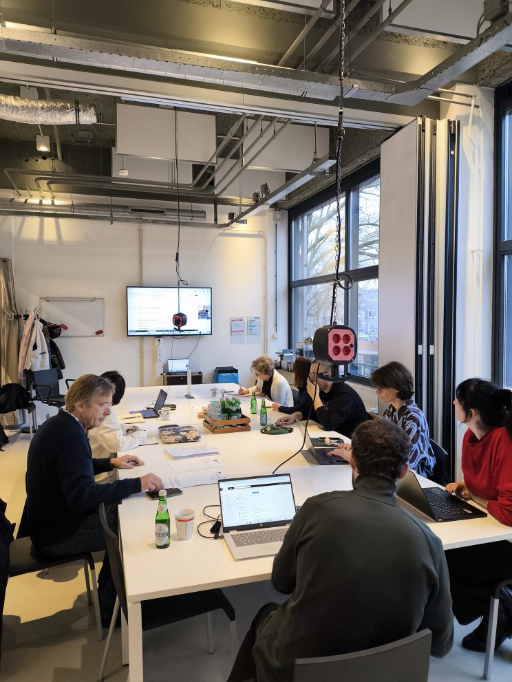 Osam osoba sjedi oko velikog pravokutnog stola u modernom radnom prostoru, svatko za svojim laptopom ili bilješkama. Na zidu je ekran s prikazanim dokumentom, a sa stropa se pruža oprema za videokonferenciju. Na stolu su boce vode, grickalice i uredski materijal, a prostorija je osvijetljena prirodnim svjetlom kroz velike prozore.