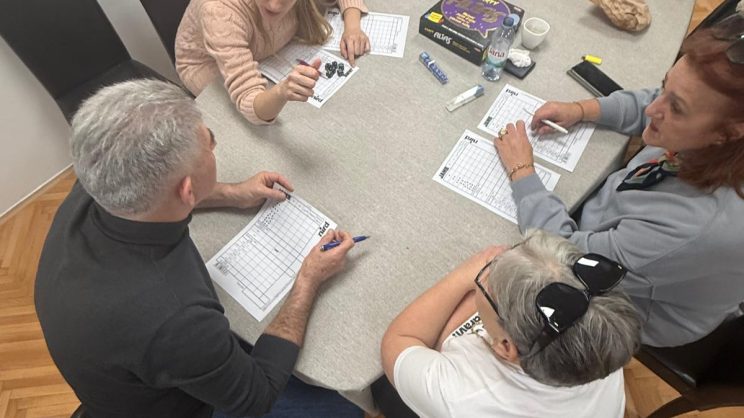 Ivana Mitar i videća pratiteljica Sanda s dvoje članova. Svi zajedno sjede za stolom i igraju društvenu igru Alias