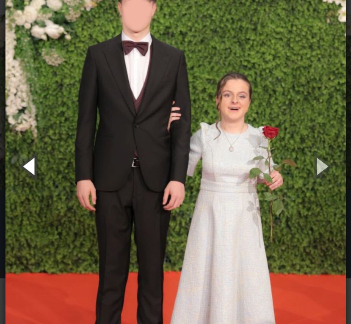 Sara pozira u bijeloj haljini s pratiteljem na crvenom tepihu, u lijevoj ruci drži crvenu ružu, a u pozadini se nalazi luk od bijelog cvijeća i zeleni zid od biljaka kao dekoracija