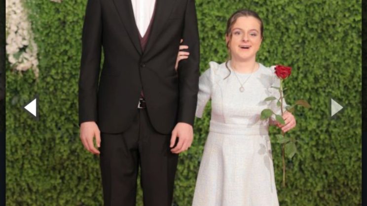 Sara pozira u bijeloj haljini s pratiteljem na crvenom tepihu, u lijevoj ruci drži crvenu ružu, a u pozadini se nalazi luk od bijelog cvijeća i zeleni zid od biljaka kao dekoracija