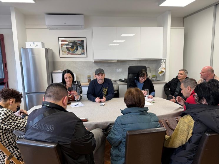 Za stolom ispred kuhinje u udruzi sjede licima okrenuti članovima s lijeva na desno: Nena Bulaja, Ivan Tokić i Jasna Giljum Kukoč. Za ostalim mjestima za stolom sjede članovi udruge koji su došli sudjelovati na Zboru članstva.