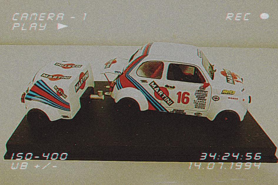 Autić s naljepnicama Martini Racing i brojem 16 vuče malu prikolicu u istom stilu, a oba su izložena na crnom postolju. Slika ima retro, zrnati efekt i grafiku koja podsjeća na snimanje kamerom.