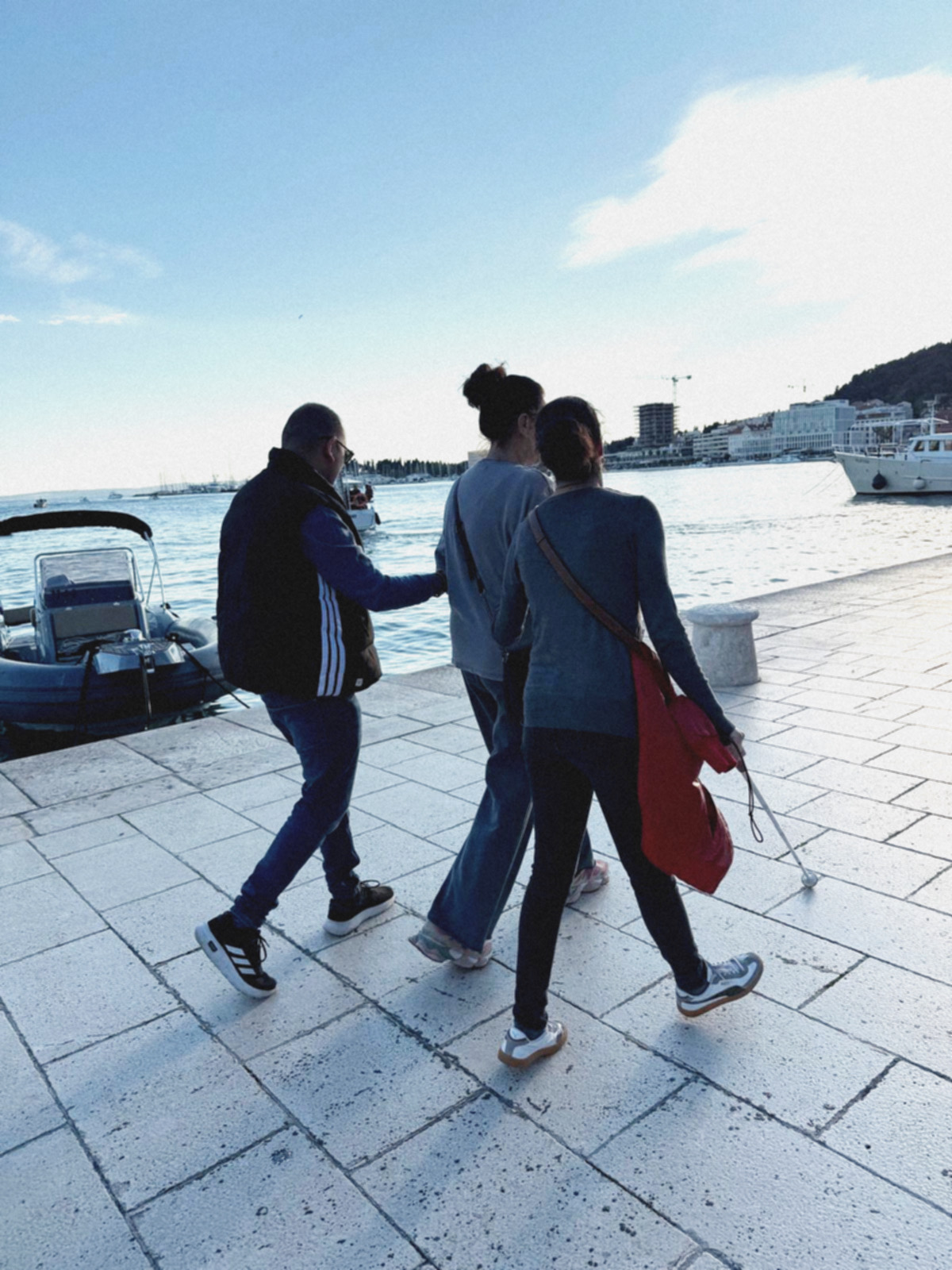 Jakov i Anđela šetaju Matejuškom uz pomoć videće pratiteljice. Na rivi je zavezan gumenjak, a u pozadini se vide brodovi i veduta Splita.