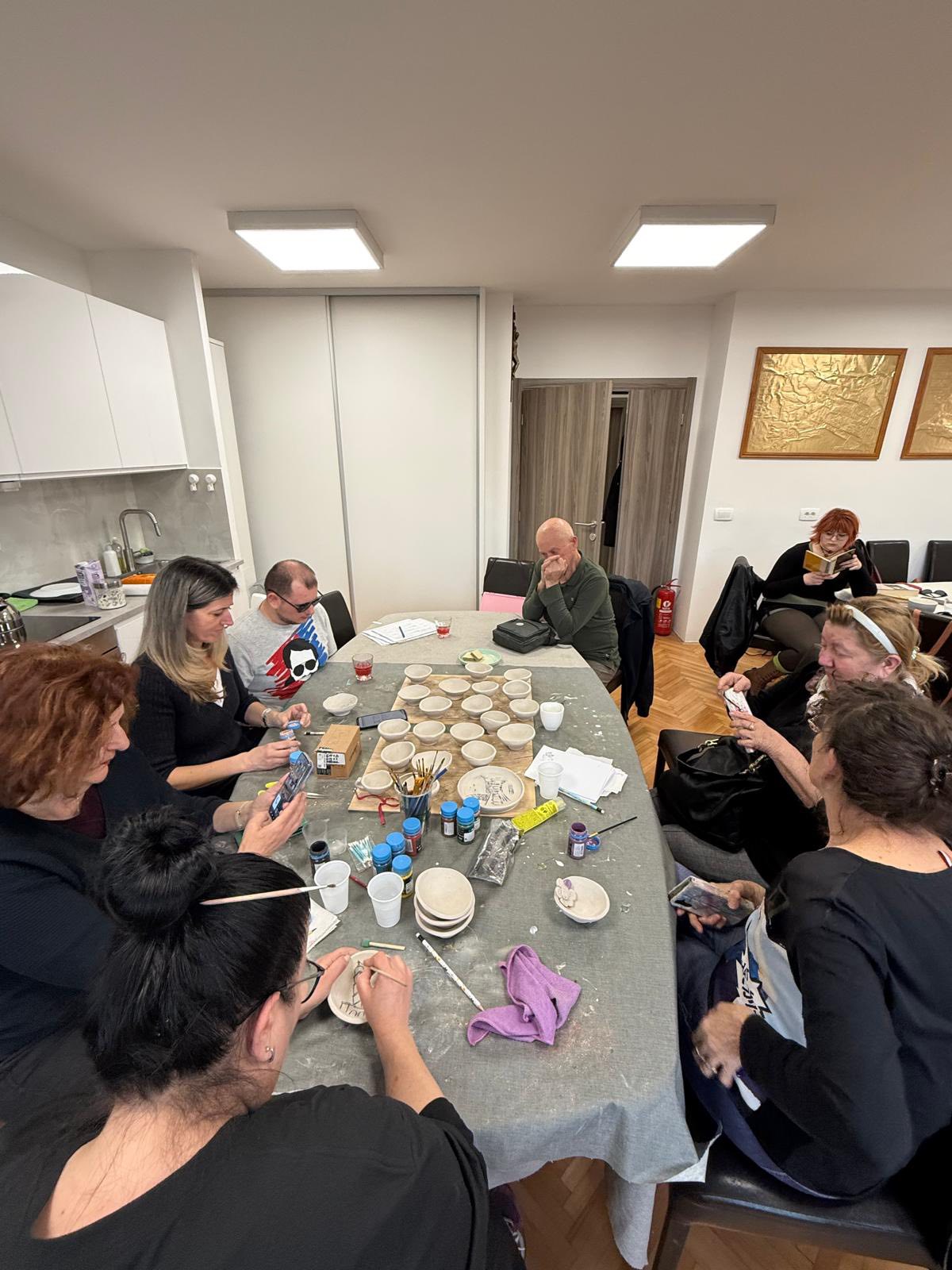 Skupina ljudi sjedi oko stola u dobro osvijetljenoj prostoriji i oslikava keramičke tanjure i zdjelice. Na stolu su razni likovni materijali i piće, a atmosfera djeluje usredotočeno i kreativno.