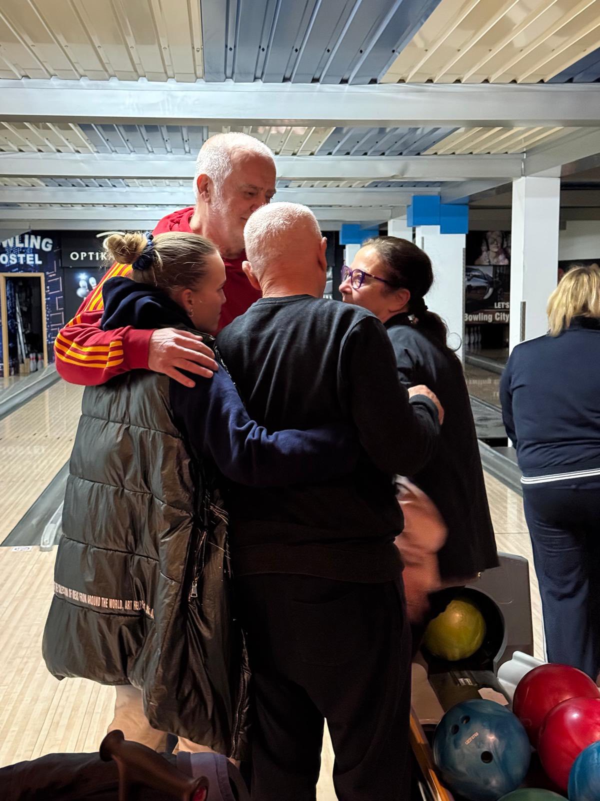 Četvero ljudi stoji zagrljeno i nasmijano u kuglani, uživajući u zajedničkom druženju pokraj stalka sa šarenim kuglama za kuglanje.