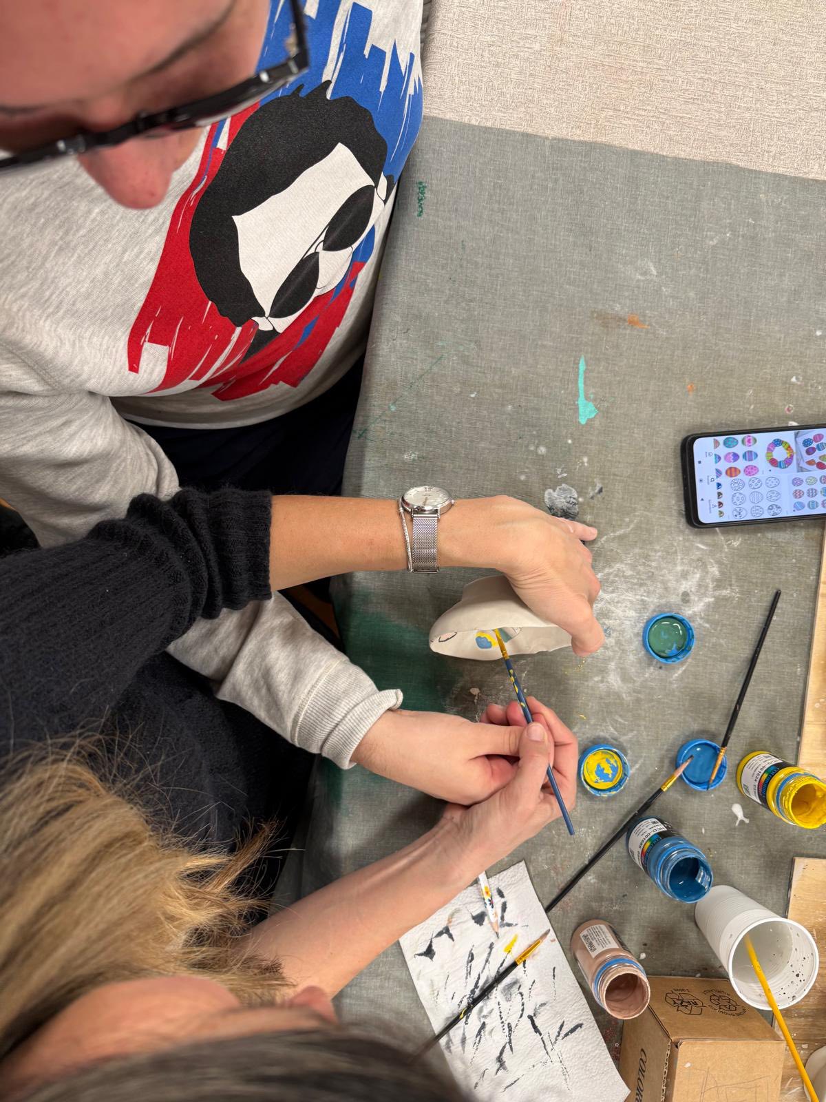 Pogled odozgo prikazuje dvije osobe kako zajedno oslikavaju keramičku posudu na stolu. U blizini se nalaze razne bočice boje, kistovi i mobitel, a na stolnjaku su vidljivi tragovi boje.