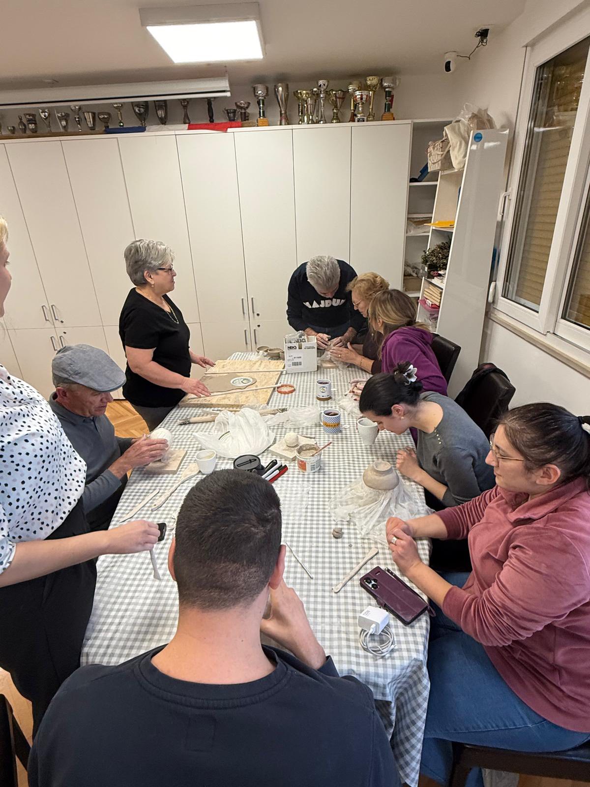 Skupina ljudi sjedi oko stola prekrivenog stolnjakom i izrađuje rukotvorine od gline i drugih likovnih materijala, dok dvije žene stoje sa strane i promatraju. Sve se odvija u prostoriji s bijelim ormarićima, a iznad njih su izloženi pehari.