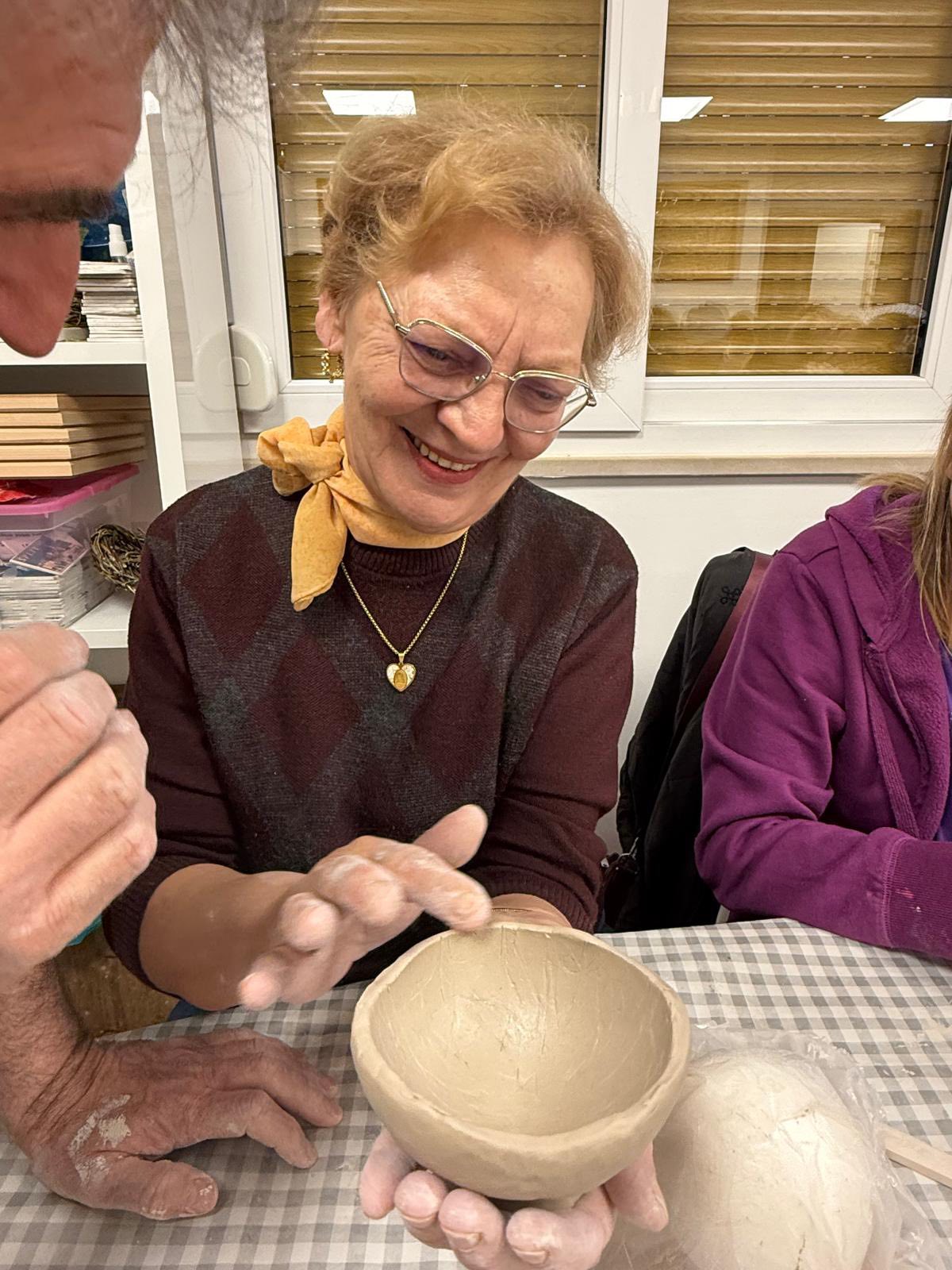 Starija žena s naočalama i žutim šalom nasmiješeno rukama oblikuje glinenu zdjelicu za stolom, uz pomoć druge osobe. Čini se da uživa u keramičarskoj radionici.