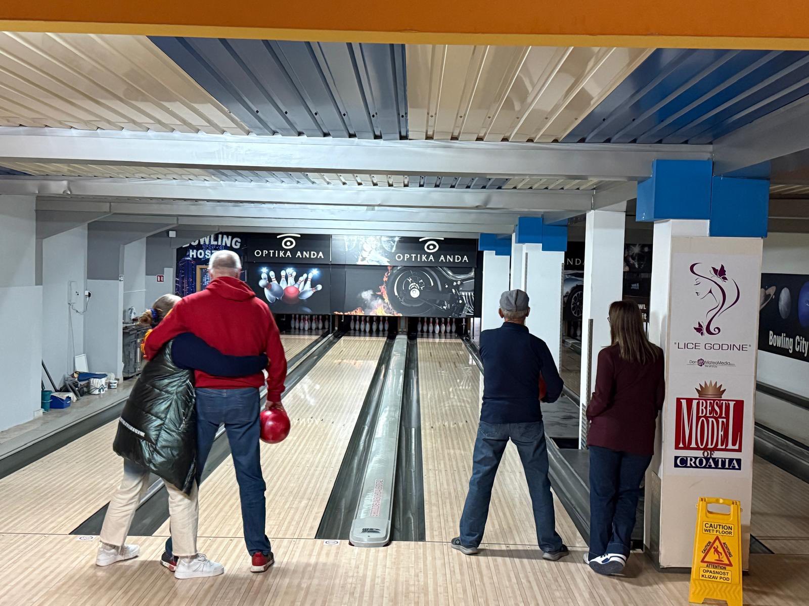 Četiri osobe nalaze se u kuglani. Dvije osobe s lijeve strane pripremaju se za bacanje kugle, pri čemu jedna grli drugu, dok dvije s desne strane promatraju staze. Na kraju staza vide se čunjevi i oznake.