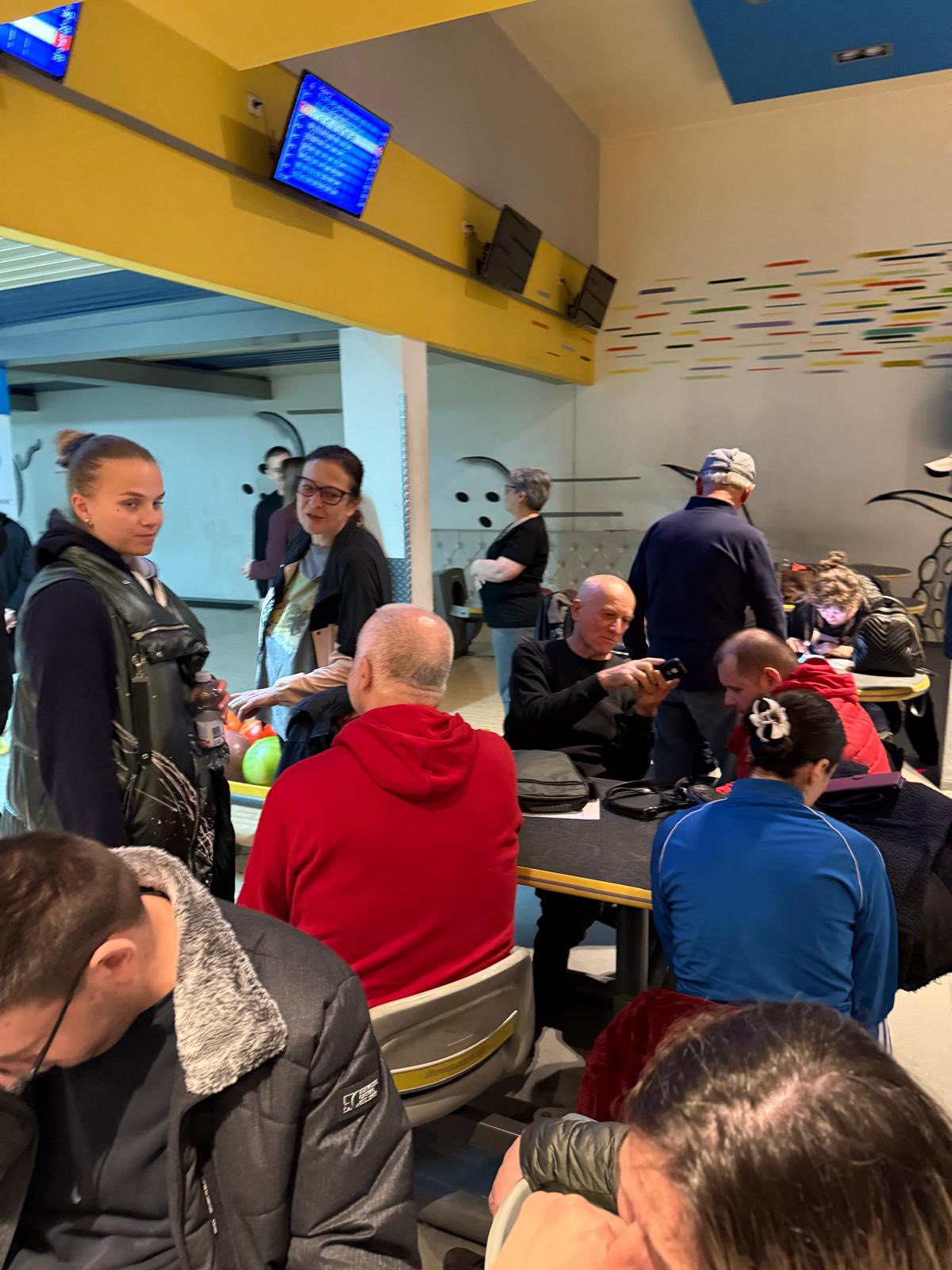 Skupina ljudi sjedi i stoji oko stolova u užurbanoj kuglani. Iznad njih vise ekrani s prikazom rezultata, a neki drže kugle za kuglanje ili međusobno razgovaraju. Atmosfera djeluje živo i društveno.