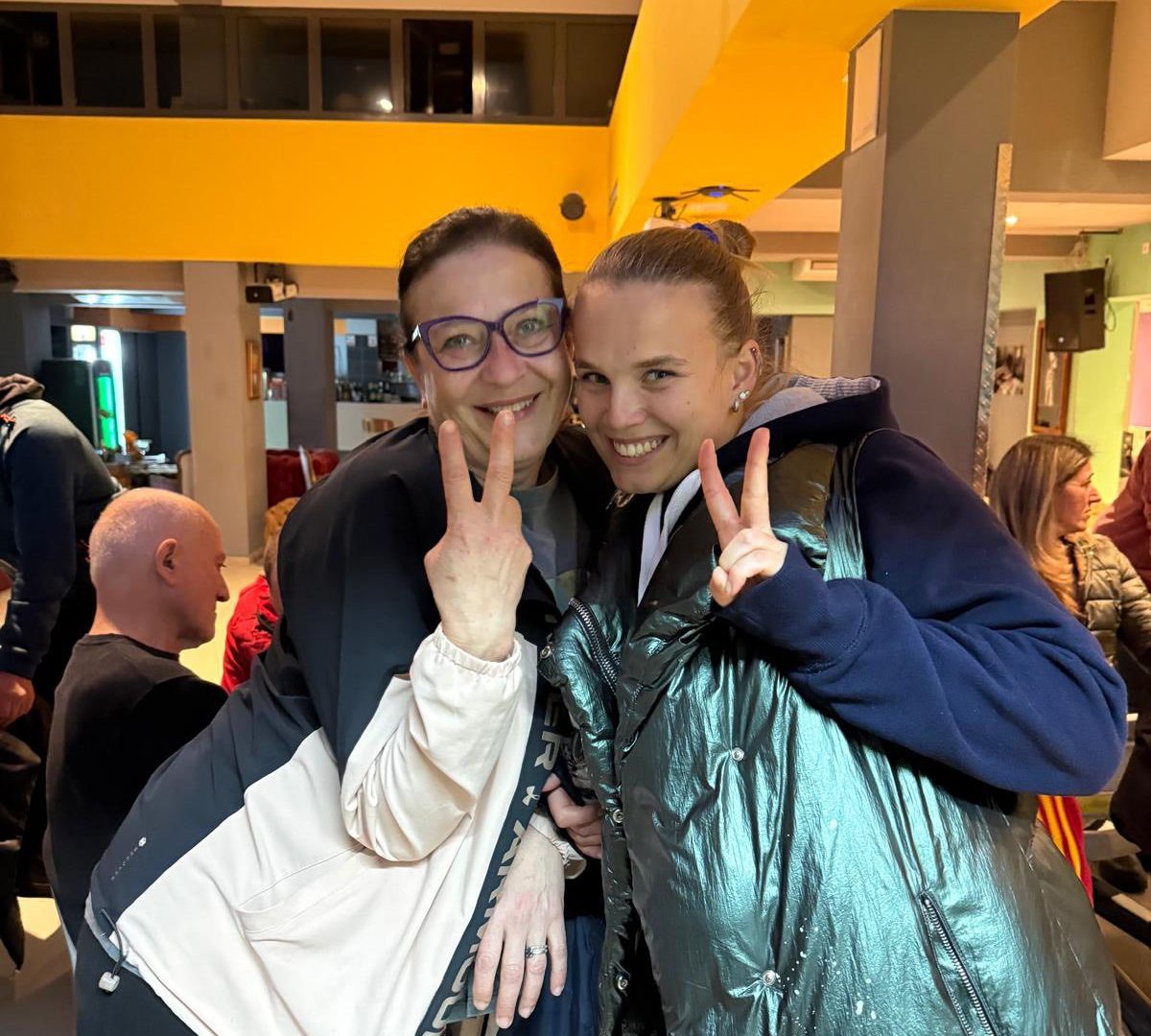 Dvije žene nasmijano poziraju u zatvorenom prostoru na živom i vedrom druženju, pokazujući znak mira prstima, dok u pozadini drugi sjede i razgovaraju.