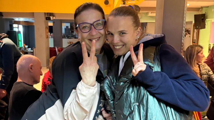 Dvije žene nasmijano poziraju u zatvorenom prostoru na živom i vedrom druženju, pokazujući znak mira prstima, dok u pozadini drugi sjede i razgovaraju.