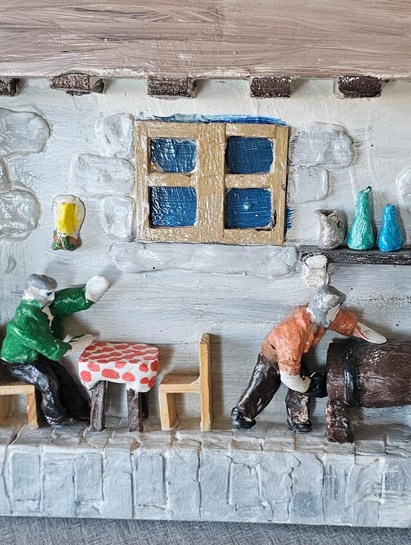 Glinena diorama prikazuje dvije osobe u rustikalnoj prostoriji s prozorom, malim stolom i dvije stolice te posuđem na polici. Jedna osoba toči iz bačve, dok druga podiže čašu.