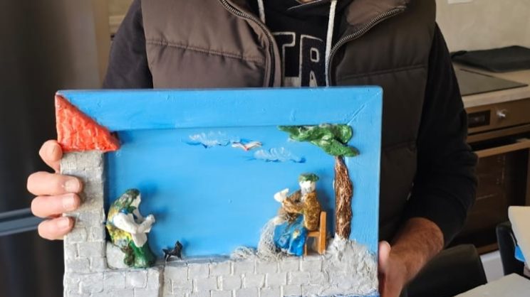 Muškarac sa sijedom kosom i bradom drži šarenu glinenu dioramu koja prikazuje dvojicu ribara uz more — jedan sjedi ispod stabla, a drugi stoji pokraj zgrade s crvenim krovom, dok se u pozadini vide plavo more i nebo.