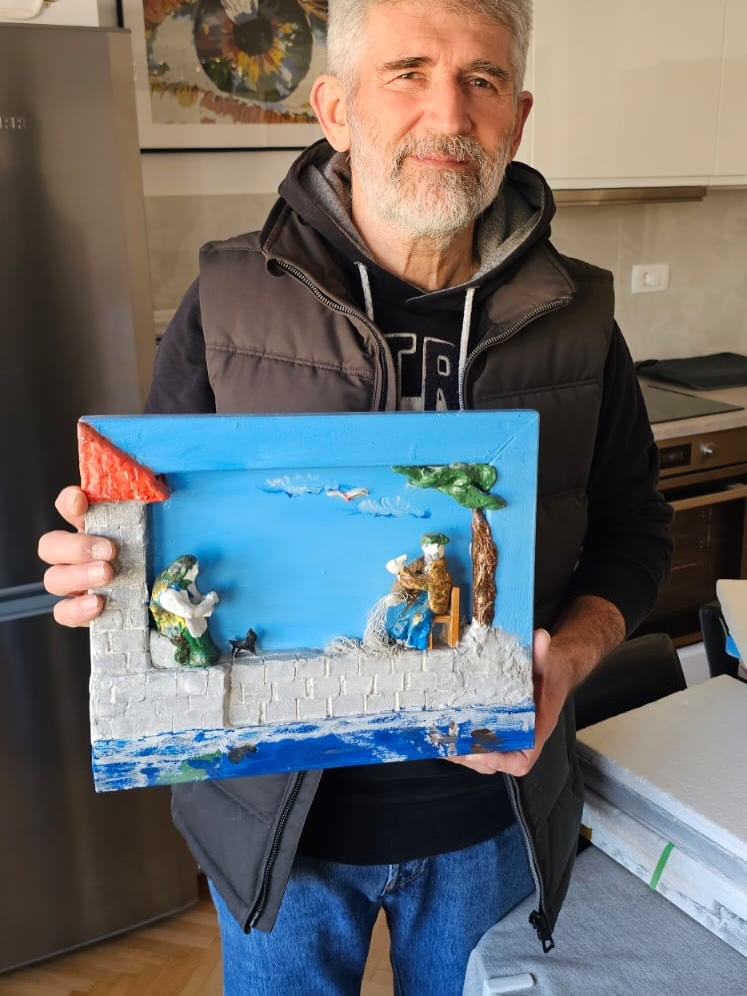 Muškarac sa sijedom kosom i bradom drži šarenu glinenu dioramu koja prikazuje dvojicu ribara uz more — jedan sjedi ispod stabla, a drugi stoji pokraj zgrade s crvenim krovom, dok se u pozadini vide plavo more i nebo.