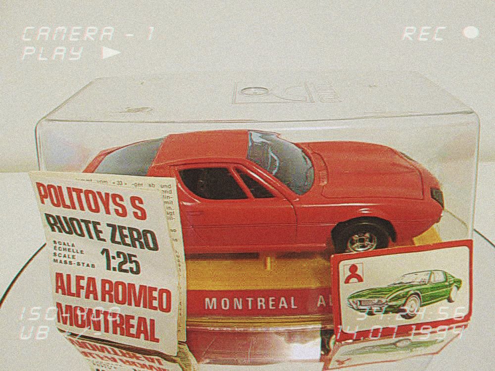 Crveni model automobila Alfa Romeo Montreal nalazi se u prozirnoj izložbenoj kutiji, s oznakom mjerila 1:25 proizvođača Politoys S. Preko kutije se vide vintage grafike, uključujući ilustraciju zelenog automobila i retro natpis, što cijeloj slici daje nostalgičan dojam stare VHS snimke.