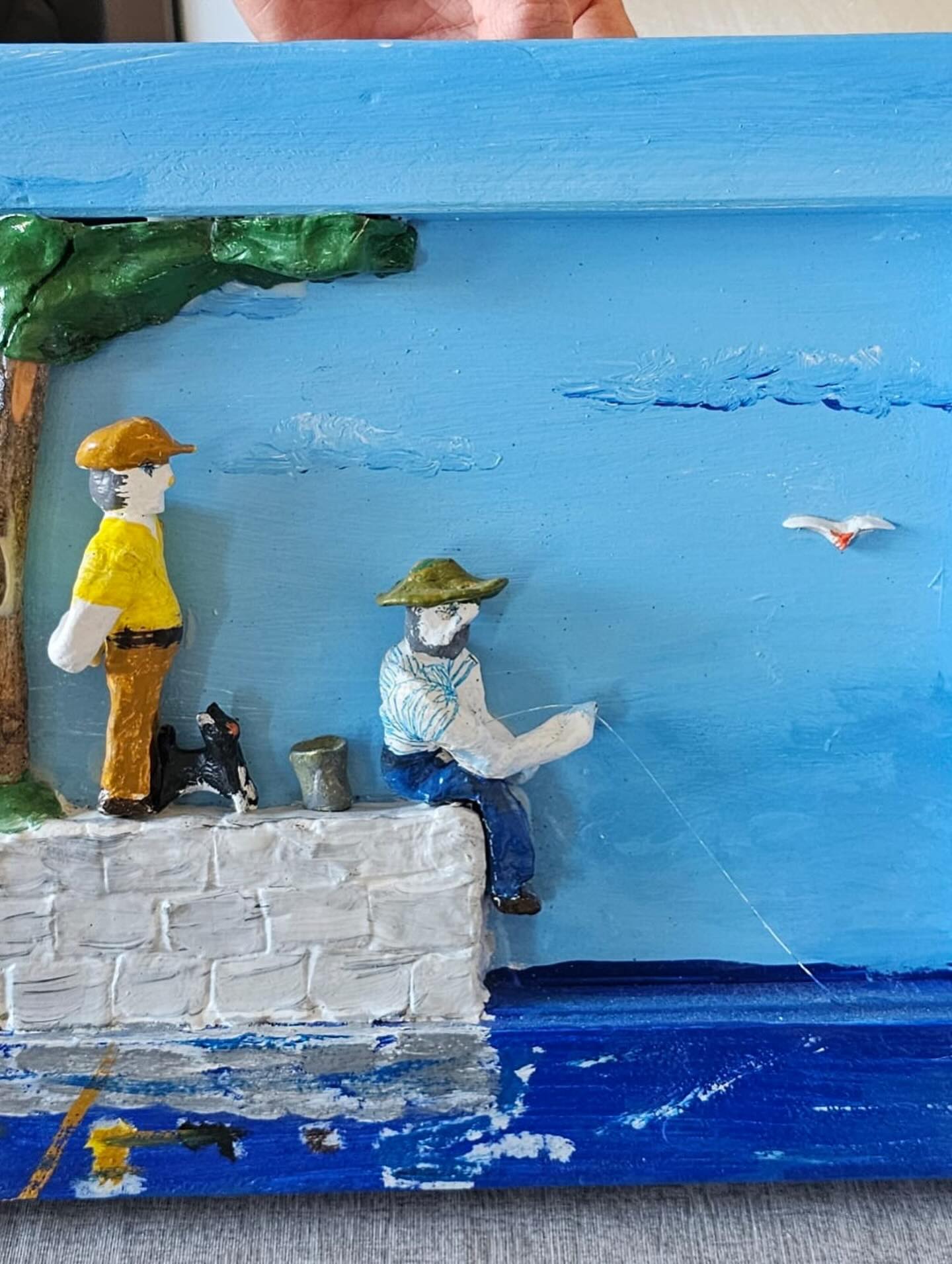 Reljefno umjetničko djelo prikazuje dvije osobe uz more — jedna stoji sa psom, dok druga sjedi i peca s bijelog kamenog ruba. U prizoru se još vide stablo, more i ptica na plavom nebu.