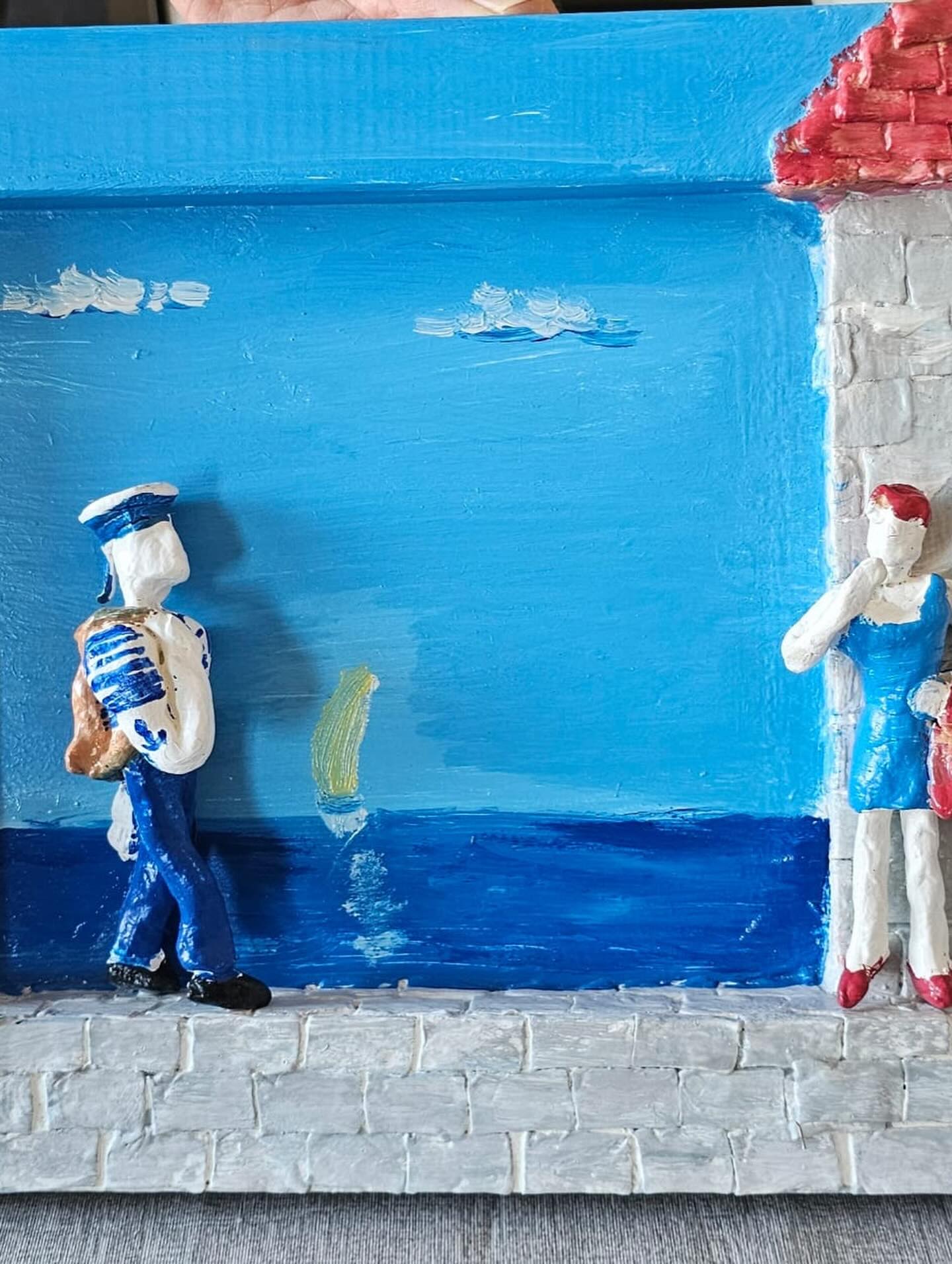 Oslikani glineni reljef prikazuje mornara kako hoda kamenim pločnikom uz more, dok žena u plavoj haljini stoji pokraj bijele kamene kuće s crvenim krovom. Obje su figure prikazane jednostavno i stilizirano.