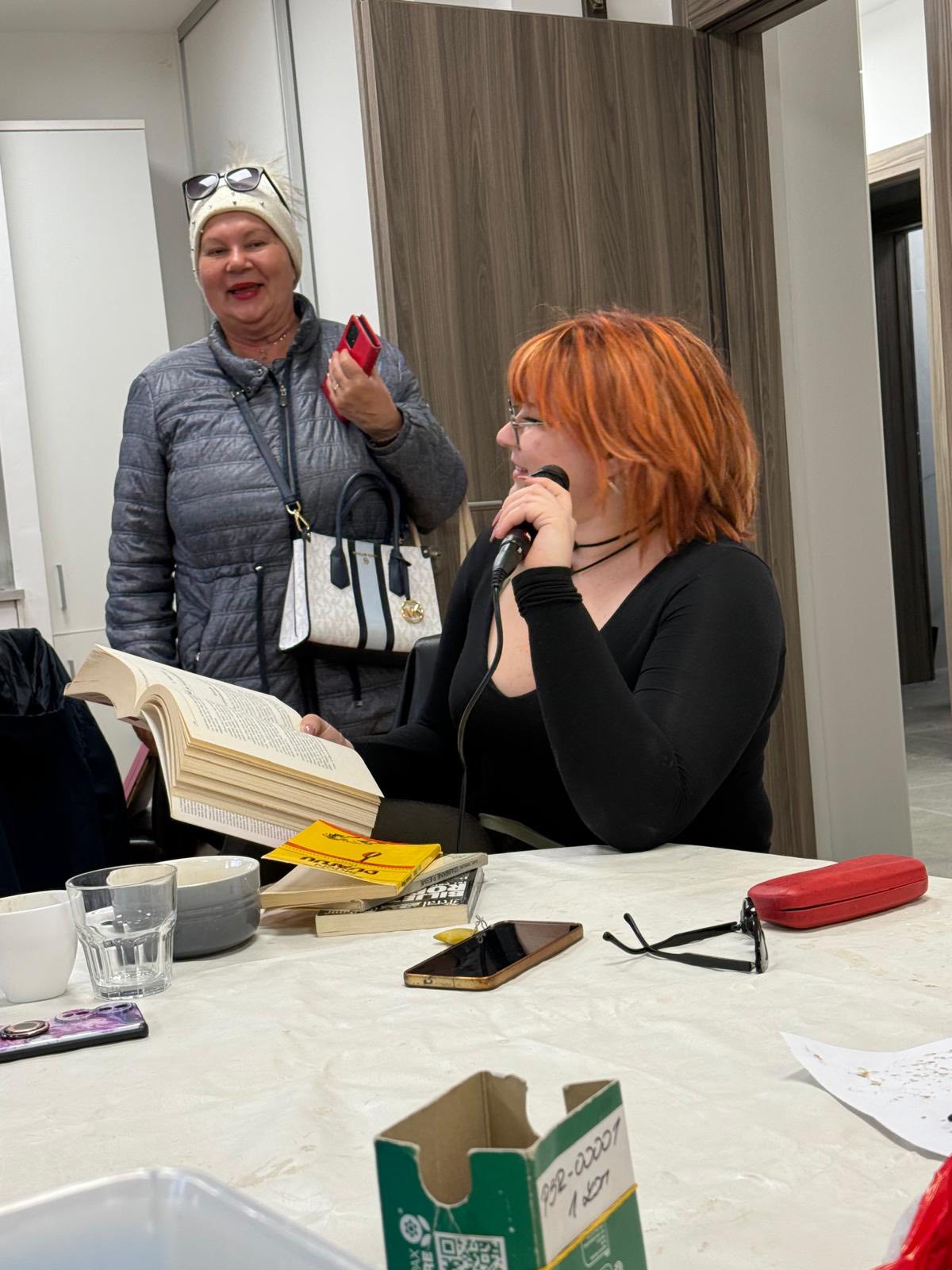 Žena crvene kose sjedi za stolom i čita iz knjige u mikrofon. U blizini stoji druga žena, u sivoj jakni i šeširu, drži mobitel i smiješi se. Na stolu se nalaze šalice, papiri i novčanik